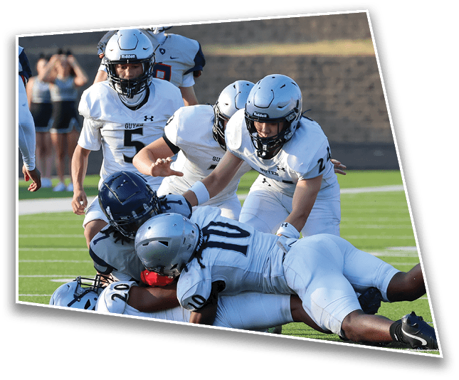 Several football players in white uniforms and helmets are tackling an opponent wearing a dark helmet on a green field during a game. Description generated by AI