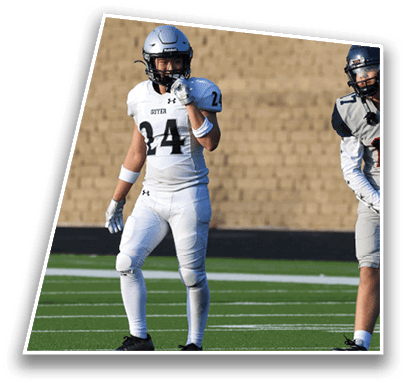 Two people wearing football uniforms and helmets stand on a green field with one person in white and the other in gray. Description generated by AI