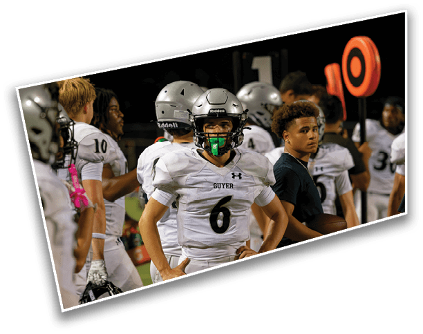 A group of people in football uniforms and helmets stand on the sidelines of a nighttime game, with one person in a black shirt among them. Description generated by AI
