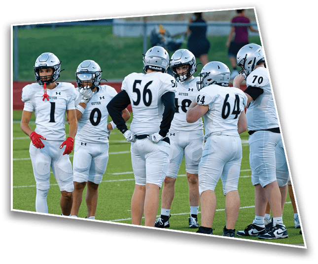 A group of people wearing white football uniforms and helmets are standing together on a green field, appearing to be in discussion or strategizing. Description generated by AI