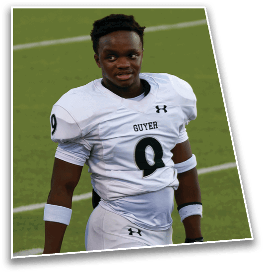 A person wearing a white football jersey with the word “GUYER" and an Under Armour logo is standing on a grassy field. Description generated by AI
