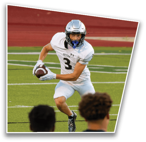 A person wearing a silver football helmet and white jersey is running on a sports field with a red track and a chain-link fence in the background. Description generated by AI