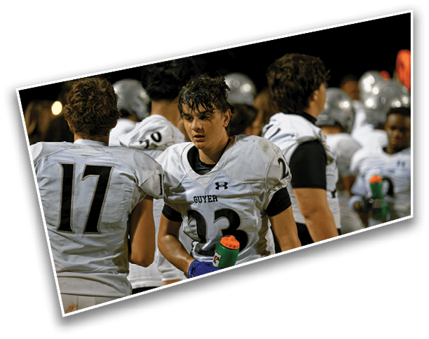 People in football uniforms are standing on the sidelines during a game, with one person in the foreground holding a water bottle and looking focused. Description generated by AI