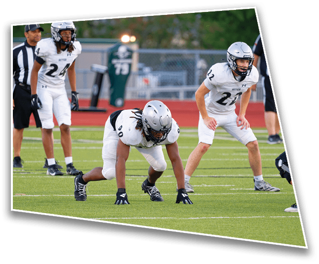 Three people in white football uniforms and helmets are positioned on a football field with one person crouched in a ready stance and two standing behind, while a referee stands in the background. Description generated by AI