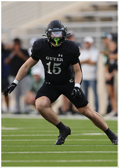 A person wearing a black football helmet and black sports uniform is looking forward on a blurred outdoor field. Description generated by AI
