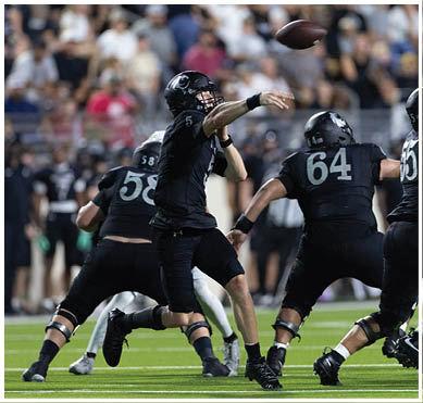 A person wearing a black football uniform is throwing a football during a game with a blurred crowd in the background. Description generated by AI