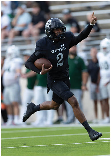A person wearing a black football uniform with the word “GUYER" and number 2 is running while holding a football on a field. Description generated by AI
