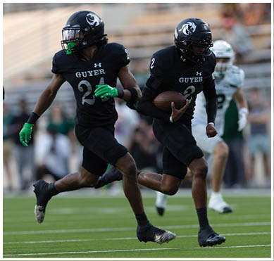 A person wearing a black football uniform with the number 24 and the word Guyer on the jersey is running on a field while holding a football. Description generated by AI