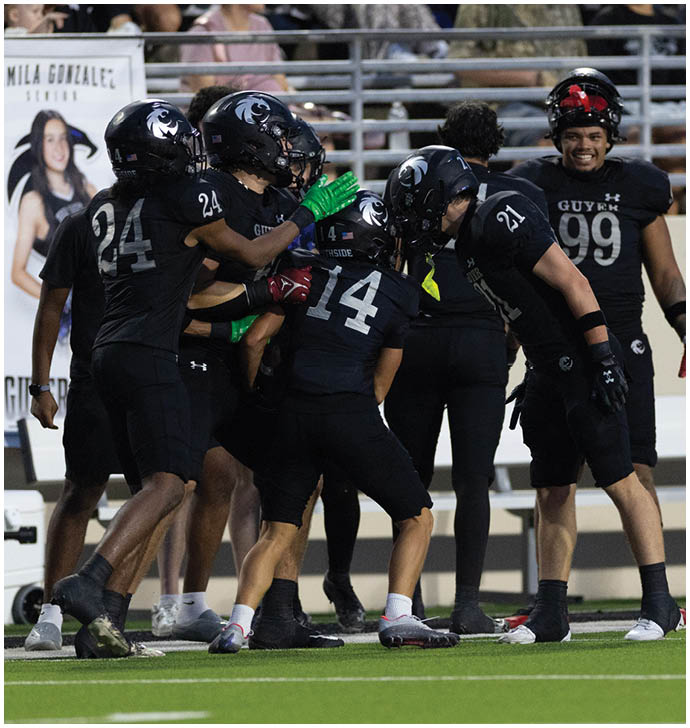 A group of people wearing black football uniforms and helmets are celebrating together on a field with spectators in the background. Description generated by AI