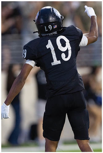 A person wearing black athletic clothing and a white glove is standing on a sports field with blurred people in the background. Description generated by AI