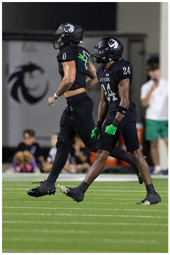 A person wearing a black sports uniform with the number zero is running on a green field with blurred people sitting in the background. Description generated by AI