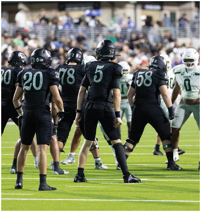 A group of people wearing black football uniforms with numbers on their backs are standing on a field facing a group of people in white and green football uniforms. Description generated by AI
