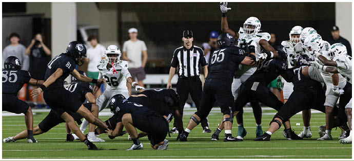 Players in dark and white football uniforms are engaged in a play on a green field with a referee observing closely in the background. Description generated by AI
