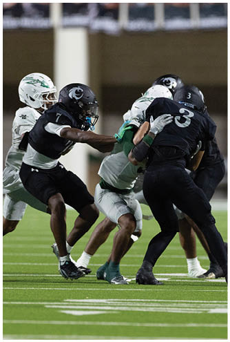 Two people in football uniforms are engaged in a play on a green field with one player in black attempting to tackle or block a player in white and green. Description generated by AI