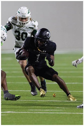 A person in black football gear with gold cleats is running with the ball while two people in white football uniforms attempt to block or tackle on a green field. Description generated by AI