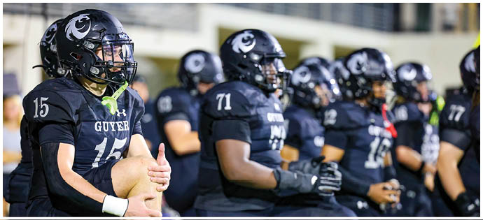 A group of people wearing black football uniforms with helmets are kneeling in a row on a field. Description generated by AI