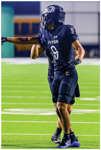 A person wearing a dark football uniform with the number 9 is standing on a field pointing to the left while another person stands behind them. Description generated by AI