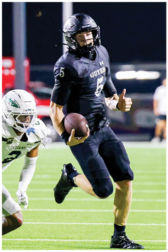 A person in a black football uniform with the number 5 is running with a football while another person in a white uniform is chasing them on a football field. Description generated by AI