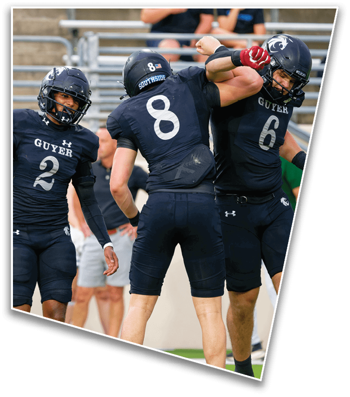 Two football players in black uniforms with helmets are on a field, one wearing number 2 and the other number 8, with people standing in the background near bleachers. Description generated by AI