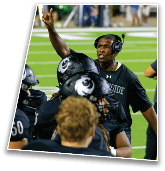 A person wearing a headset and a black shirt is animatedly gesturing and speaking to a group of people in black football helmets on a green field. Description generated by AI