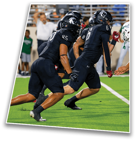 Two football players in black uniforms are running toward two players in white uniforms on a green field with spectators in the background. Description generated by AI