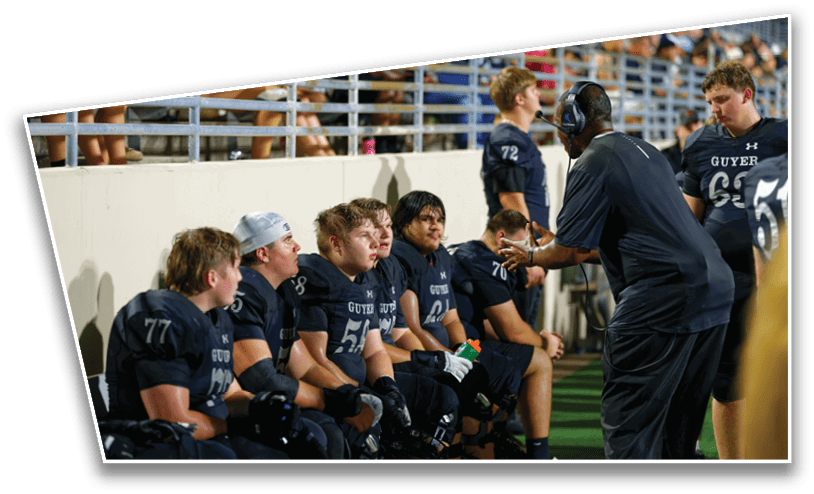 A group of people wearing football uniforms sit on a bench while a person in a headset and dark clothing talks to them on the sidelines of a sports field. Description generated by AI