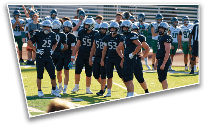 A group of people wearing dark football uniforms and helmets stand together on a field while another group in lighter uniforms and a referee stand in the background. Description generated by AI
