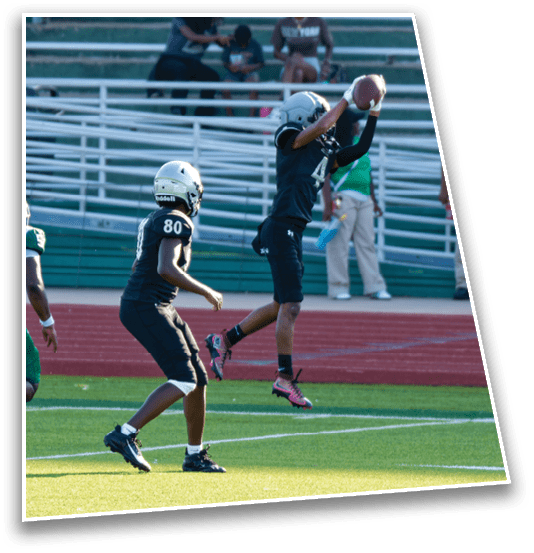 Two people in black football uniforms and helmets are on a field with one jumping to catch a football while the other watches. Description generated by AI