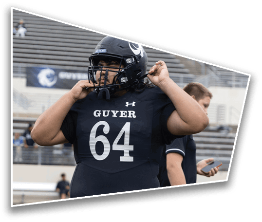 A person wearing a black football uniform with the number 64 and the word Guyer is adjusting their helmet on a football field with empty bleachers in the background. Description generated by AI