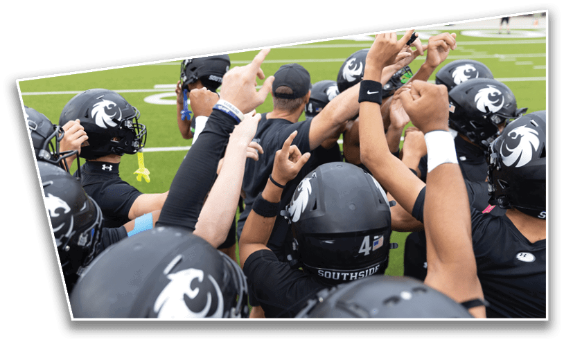 A group of people wearing black football helmets and uniforms are huddled together with their arms raised in a team gesture on a green field. Description generated by AI