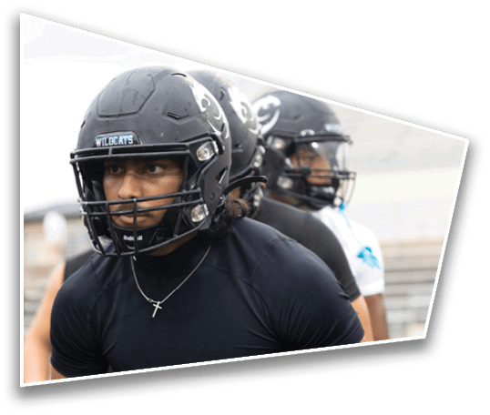 People wearing football helmets and athletic clothing are standing on a field, appearing focused and ready for practice or a game. Description generated by AI