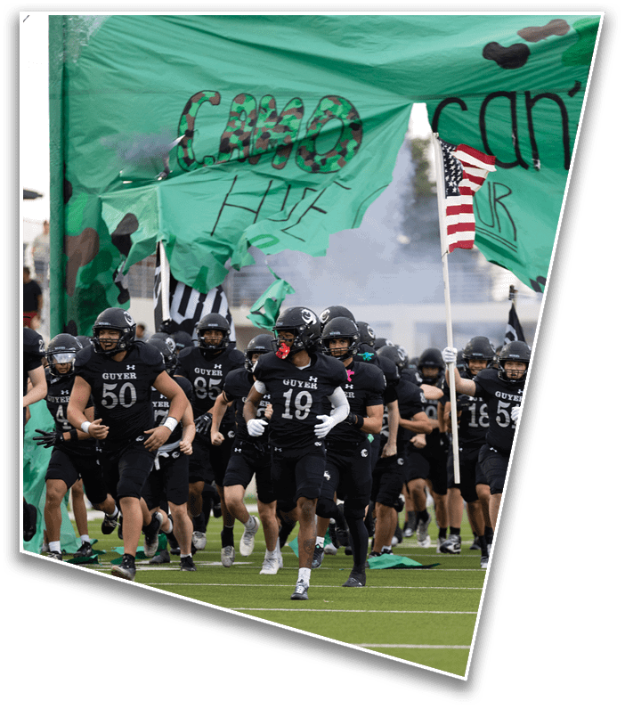 A group of people in black football uniforms and helmets run onto a field through a large green banner with camouflage patterns and partially visible text. Description generated by AI