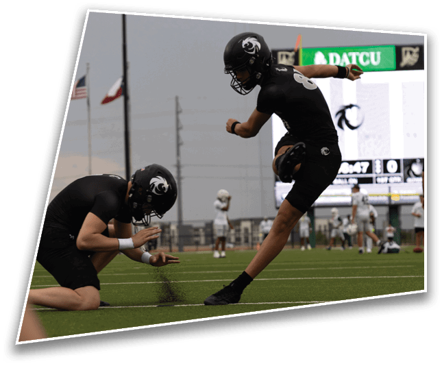Two people in black sports uniforms and helmets are on a football field with one holding the ball on the ground while the other kicks it. Description generated by AI