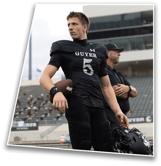 A person in a black football uniform holding a helmet and a football stands on a field with another person in the background near stadium seating. Description generated by AI