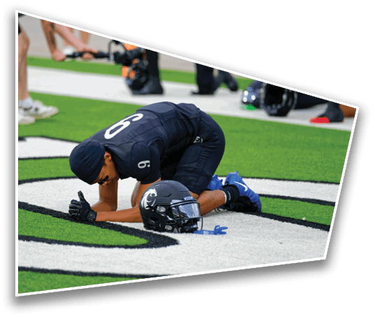 A person in a dark sports uniform kneels on a football field with their head bowed near a helmet on the ground. Description generated by AI