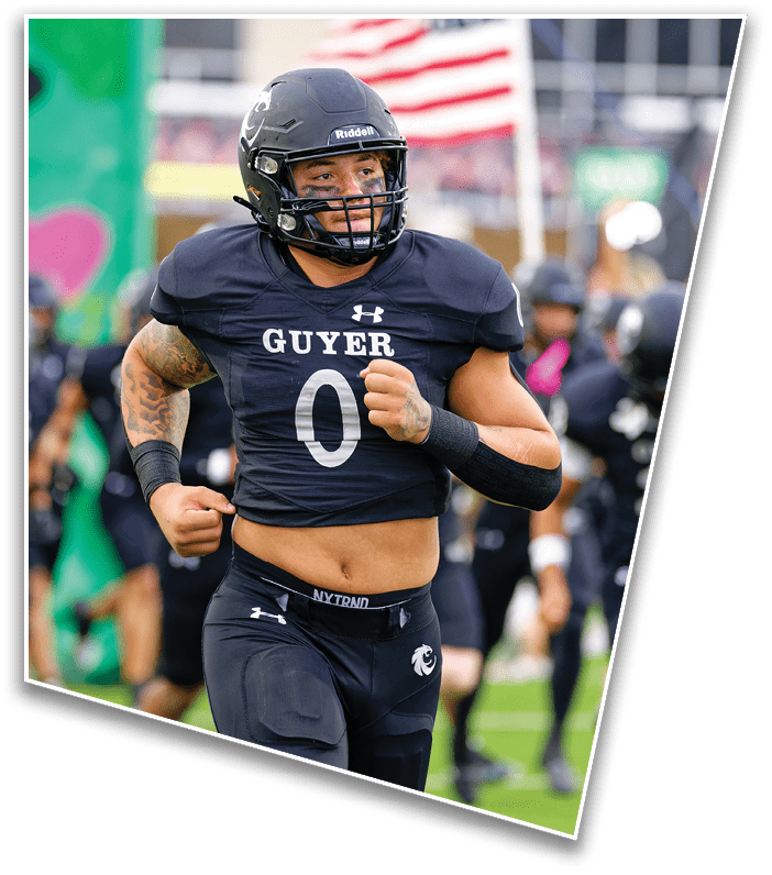A person wearing a black football uniform with the word “GUYER" and the number zero is running on a field with an American flag in the background. Description generated by AI