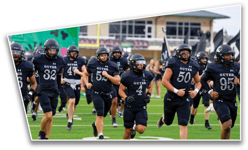 People wearing black football uniforms with helmets are running on a green field toward the camera with a stadium and spectators in the background. Description generated by AI