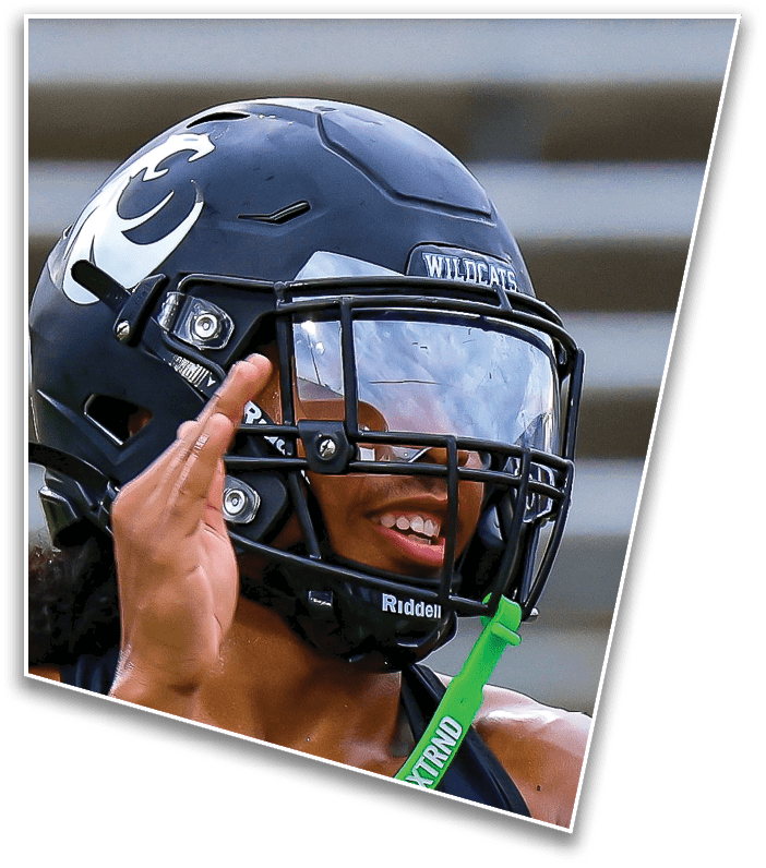A person wearing a dark football helmet with a clear visor and a green mouthguard is smiling and raising one hand. Description generated by AI