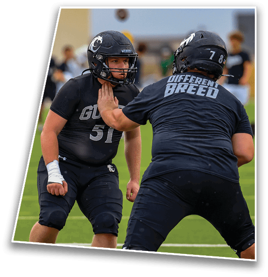 Two people wearing black football uniforms and helmets are engaged in a practice drill on a green field, with one person placing a hand on the other's chest. Description generated by AI