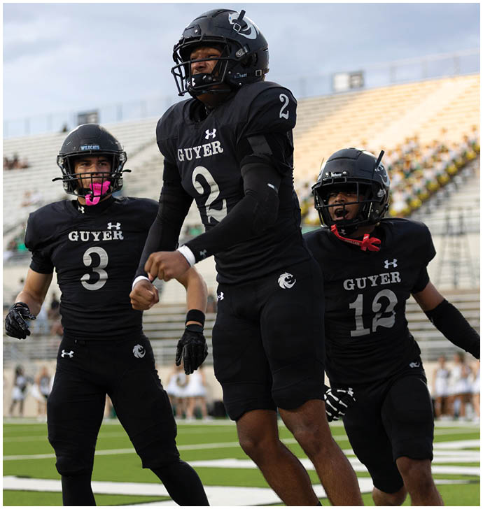 Three people in black football uniforms with helmets are on a field with bleachers in the background, with one person jumping or celebrating. Description generated by AI