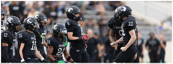 A group of people wearing black football uniforms with helmets are standing and interacting on a field during a game or practice. Description generated by AI