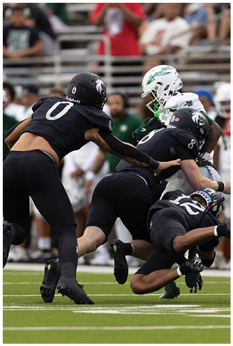 A person wearing a black helmet with a white logo is in the foreground with blurred people sitting on bleachers in the background. Description generated by AI