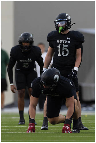 Two people wearing black football uniforms with helmets and the word “GUYER" on their jerseys are on a field with a blurred background. Description generated by AI