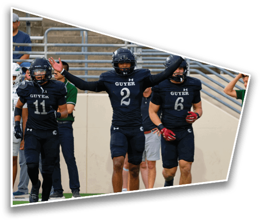 Two people in black football uniforms with helmets are walking on a field while a person in a green shirt and white pants stands nearby with a hand on their head. Description generated by AI