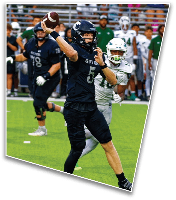 A person in a black football uniform is preparing to throw a football while being closely pursued by a person in a white uniform on a football field with spectators in the background. Description generated by AI