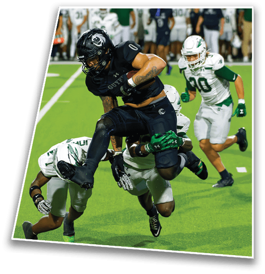 People in football uniforms are engaged in a dynamic play on a green field with one person leaping over others attempting to tackle. Description generated by AI