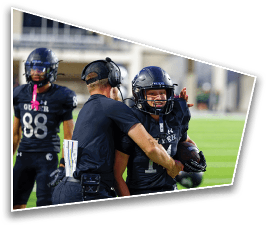 A person wearing a headset and black clothing is interacting with a football player in black uniform and helmet on a sports field. Description generated by AI