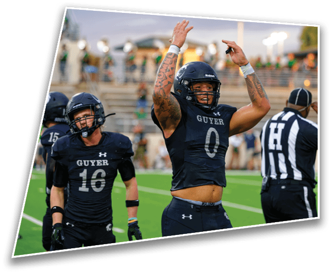 Two athletes in black football uniforms celebrating on a field with a referee nearby and blurred spectators in the background. Description generated by AI
