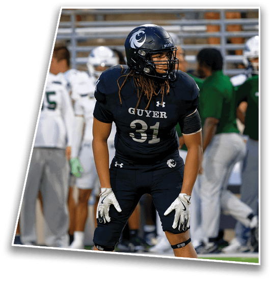 A person wearing a dark football uniform and helmet with the word “GUYER" and number 21 stands on a field with blurred players in white uniforms in the background. Description generated by AI