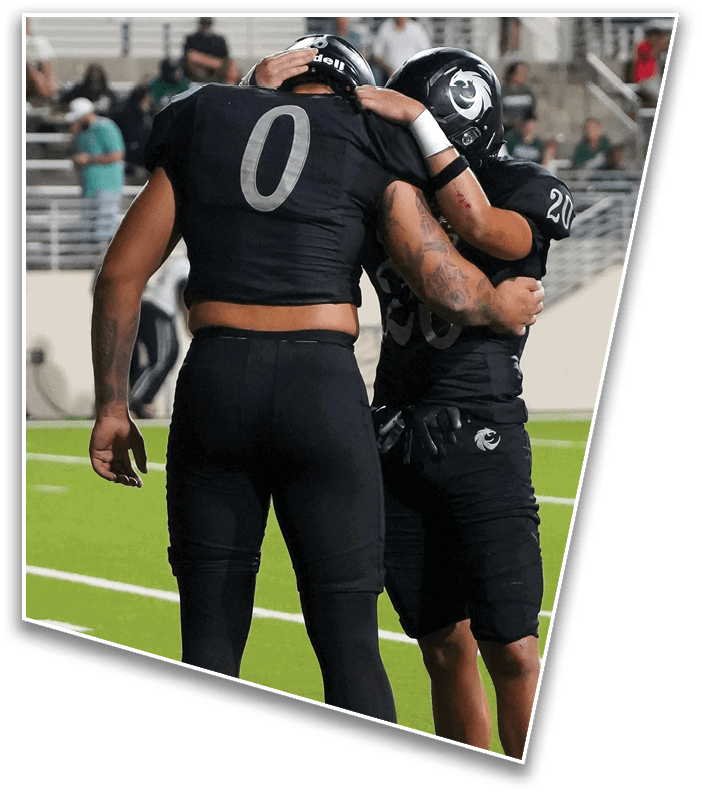 Two people wearing black football uniforms and helmets embrace on a field, with one person’s hand resting on the other’s helmet. Description generated by AI